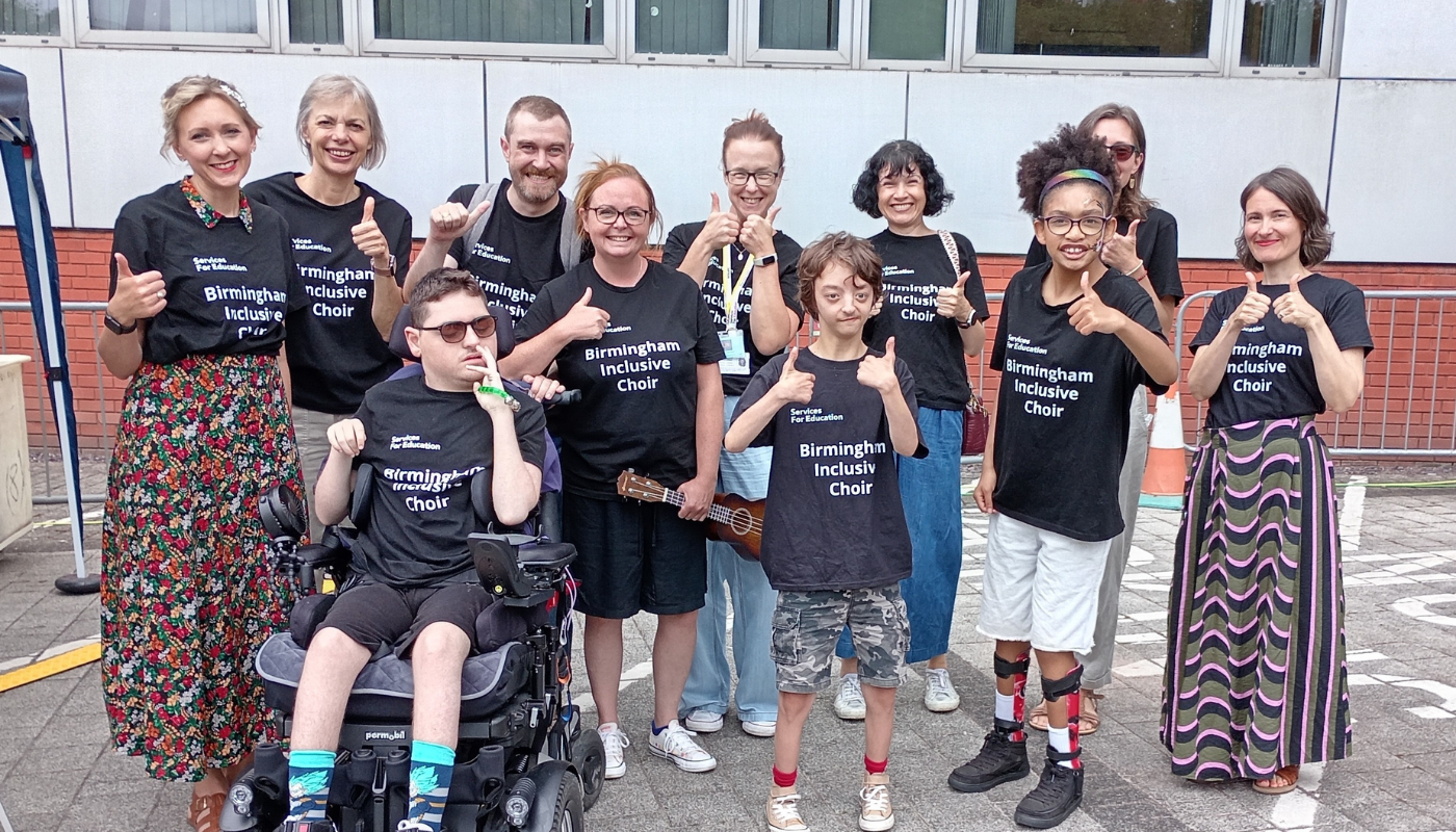 A diverse group of eleven children and adults standing outdoors on a paved area, all wearing matching black t-shirts that read "Birmingham Inclusive Choir" in white text. Everyone is smiling and posing with a thumbs-up gesture. One young man in the front sits in a motorized wheelchair, another young person holds a ukulele, and the group displays a warm, joyful community atmosphere.