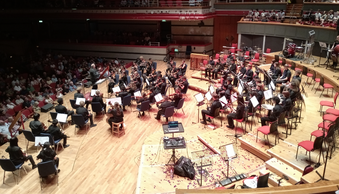 A wide-angle shot of Services For Education's Music Ensembles performing at Symphony Hall in Birmingham, a multi-tiered concert hall filled with an audience. The musicians, dressed in dark attire, are seated with violins, cellos, brass, and percussion instruments surrounding a conductor. Bright red confetti is scattered across the wooden floor around a DJ setup and audio equipment near the front of the stage.