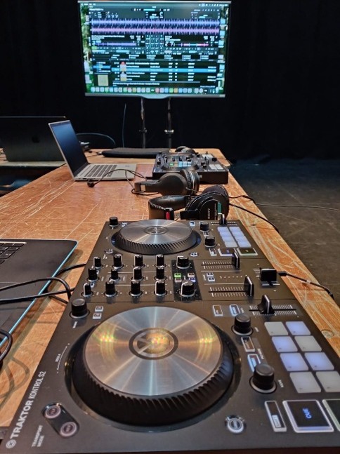 A close-up view of a DJ controller set up on a wooden desk, surrounded by headphones, laptops, and a large monitor displaying DJ software with audio waveforms.
