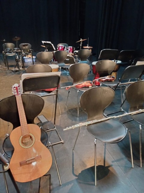 An empty rehearsal space set up with rows of black chairs holding various musical instruments, including a classical guitar in the foreground, a flute, violins, and a drum set in the background against a dark curtain.